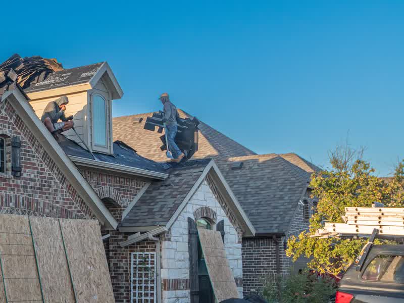 Two roofers stripping old shingles off a brick house, plywood and ladders staged in the yard