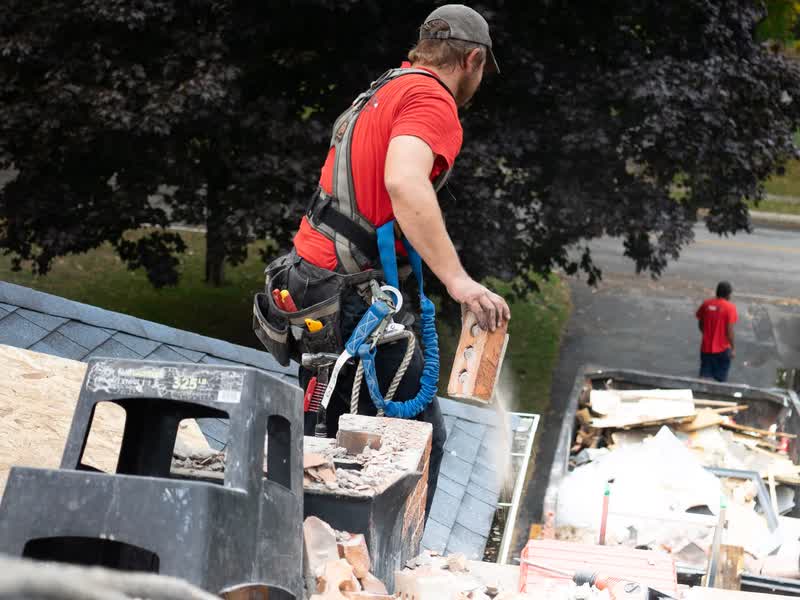 A roofer in a safety harness dropping debris into a job-site dumpster while a crewmate works below